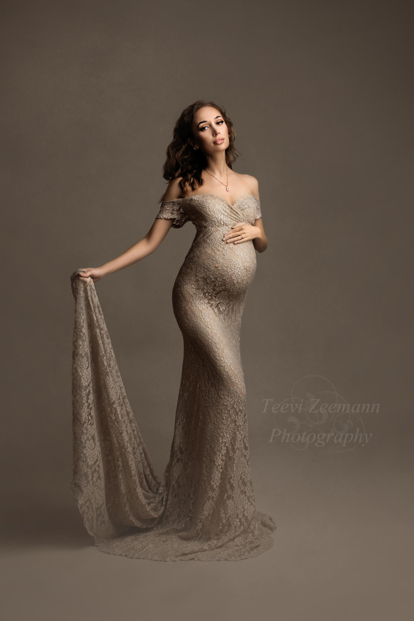 pregnant woman poses wearing a tight long dress made of lace in sand color. the top is off shoulder, with short sleeves and an adjustable sweetheart neckline. she is facing the camera, holding her bump with one hand and holding the train of the dress with the other one.