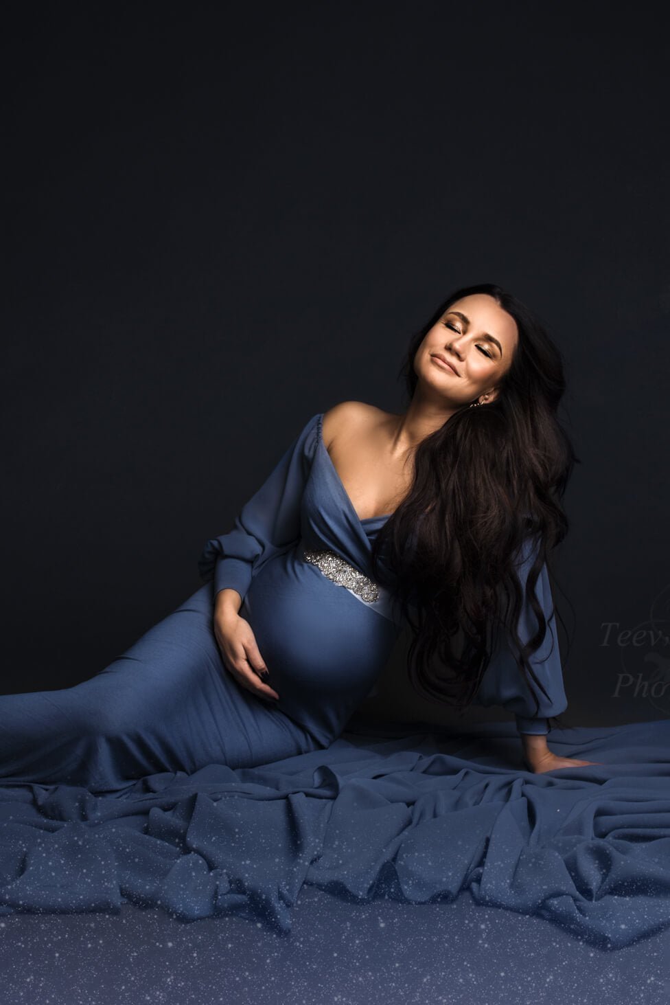 brunette model poses with a denim long maternity dress and a sash to match the style. the model is sitting on the floor, looking above and has her eyes closed.