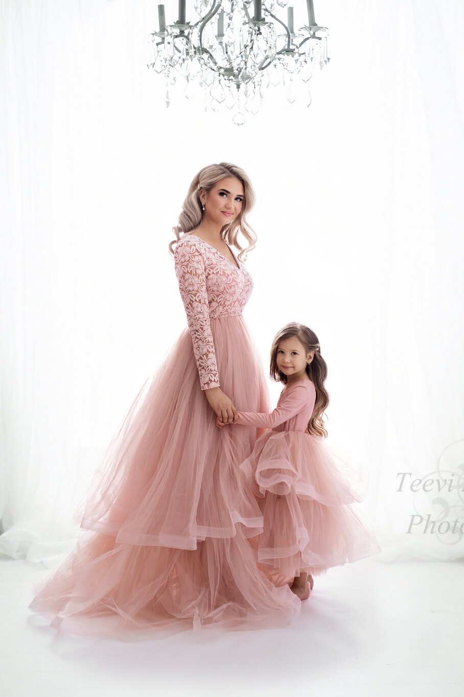 A mother is holding the hand of her little daughter. They both have a pink dress with a tulle skirt on. they are looking at the camera 
