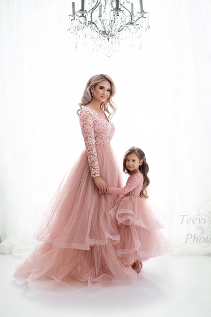 A mother is holding the hand of her little daughter. They both have a pink dress with a tulle skirt on. they are looking at the camera 