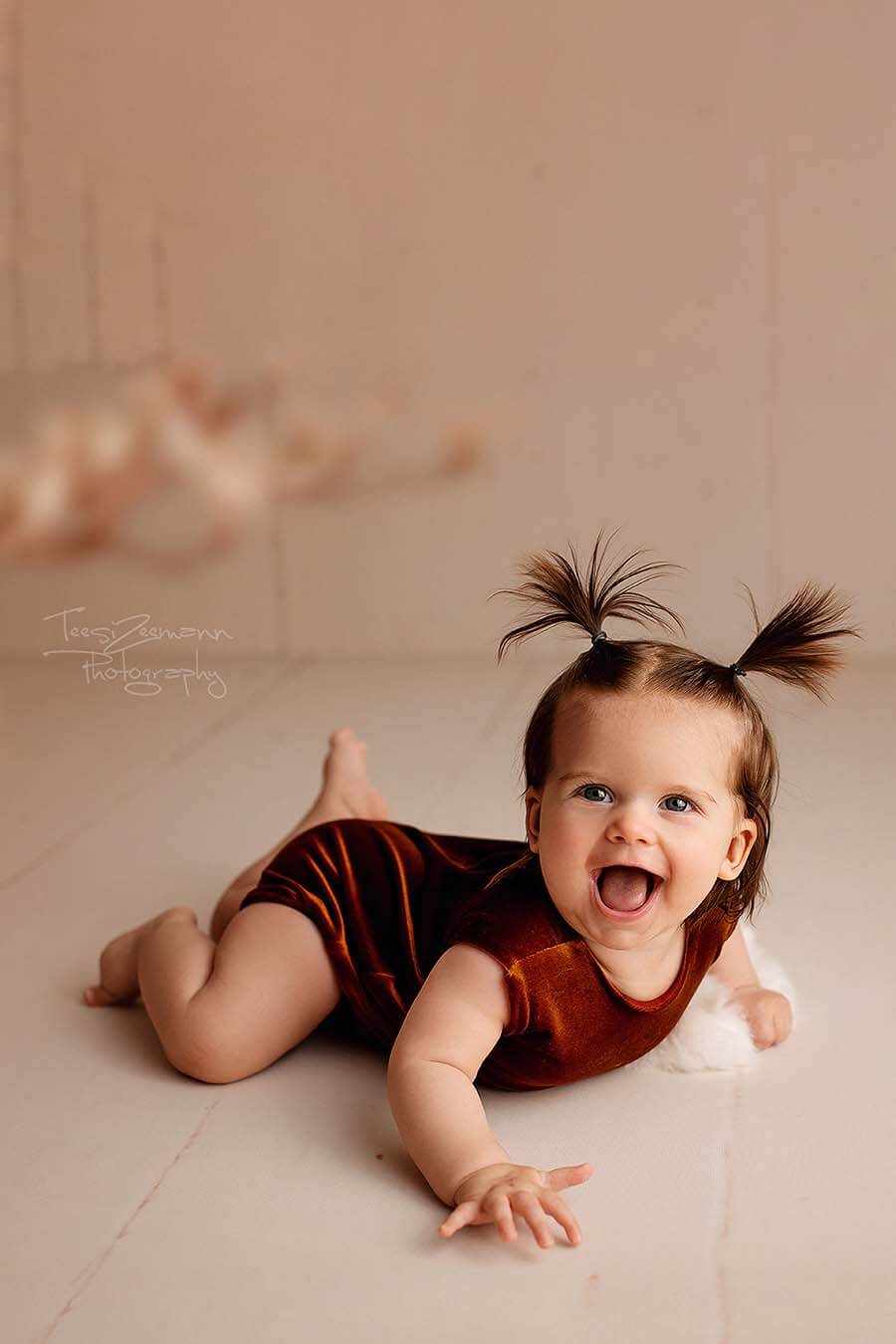 A little baby girl is laying on the ground. She has two ponytails in her hair. She is wearing a velour romper