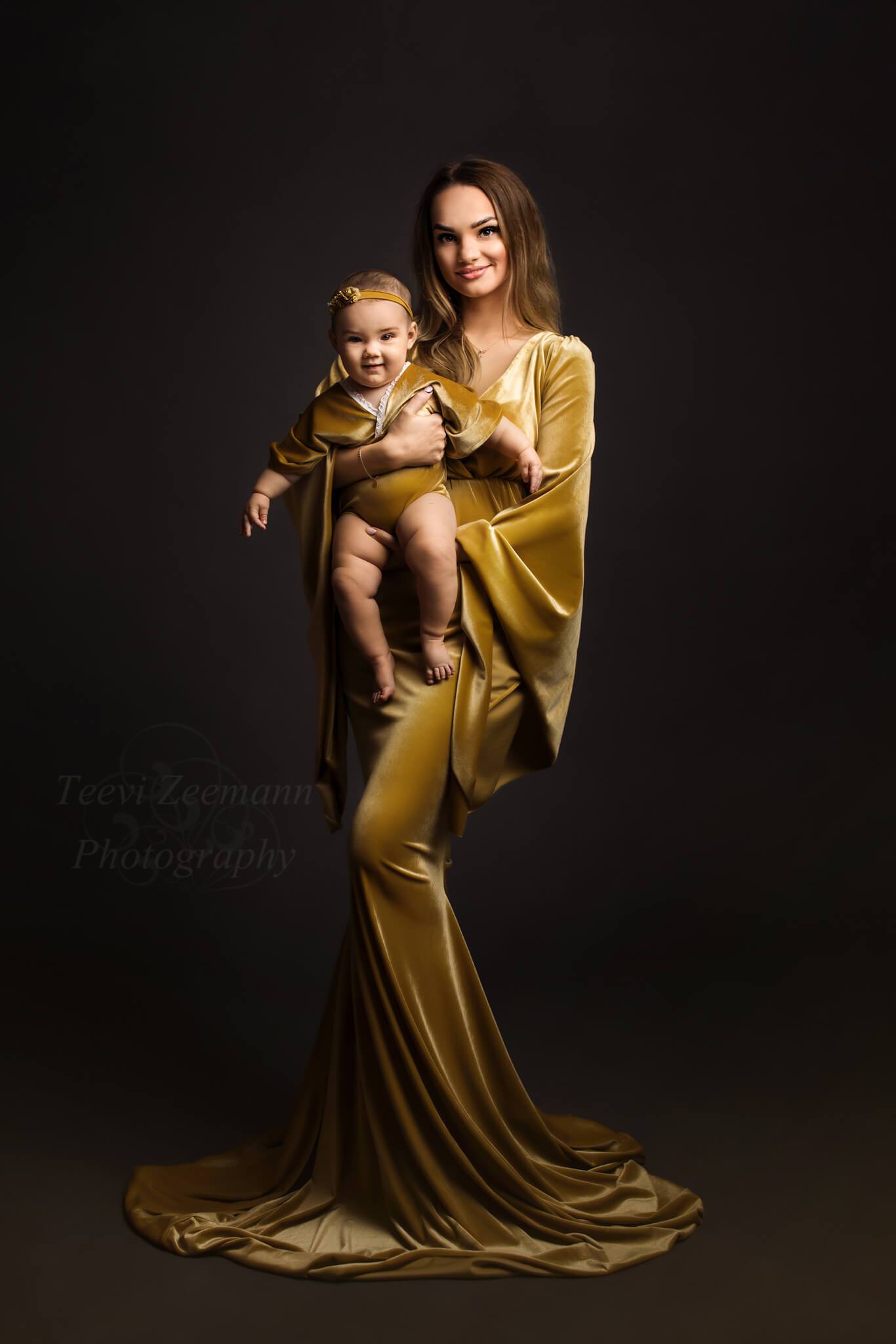 A gorgeous mother is holding her baby. They are both wearing a velour outfit in the color amber. The mother wears a long dress with boho sleeves . The baby wears a romper and has a little headband on