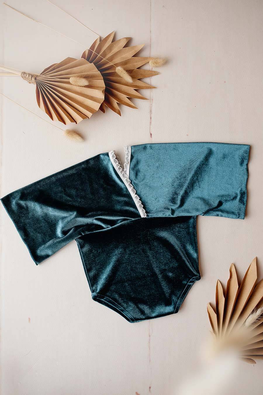 A newborn romper in a blue color. The romper is made from velour and has en little lace line at the neck. The romper is laying on the ground
