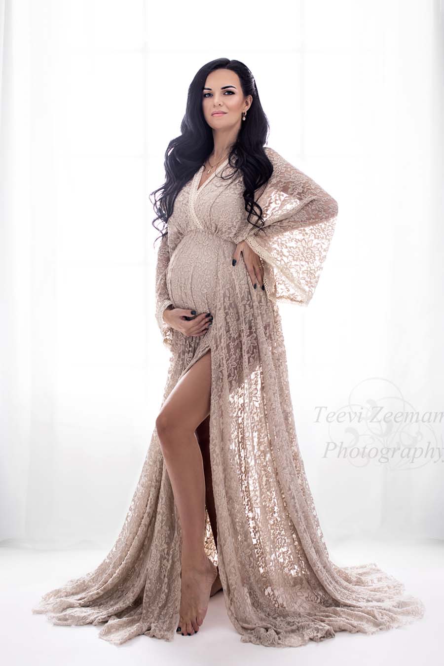 dark haired pregnant model poses in a studio wearing a long dress made of lace in sand color. she stares at the camera while holding her waist and bump. the top of the dress has a low v cut neckline and kaftan sleeves. the long circle skirt has a side split.
