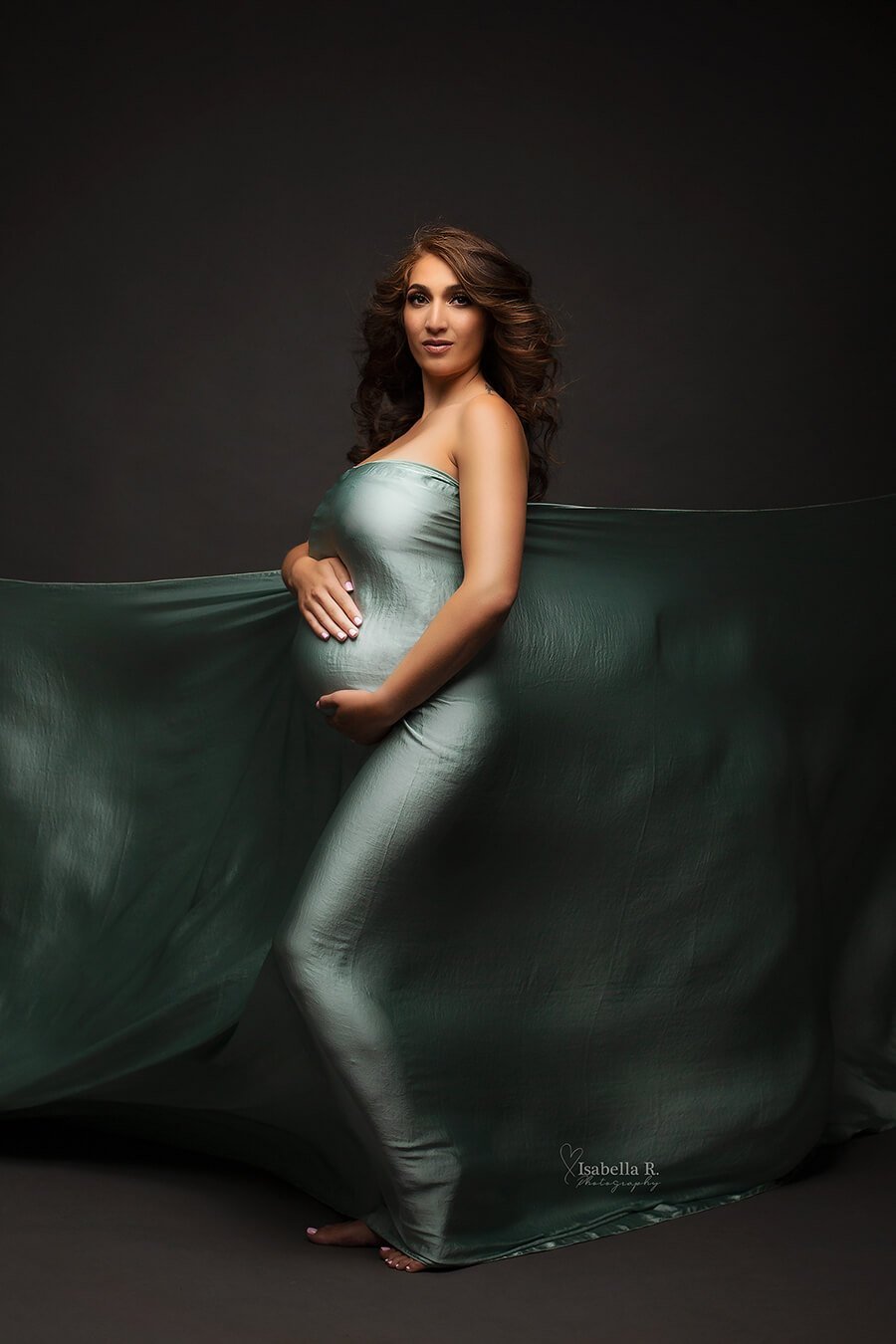 A pregnant woman is wrapped in a silky scarf. The scarf is in the colour azur. She has brown short hair and looking towards the camera