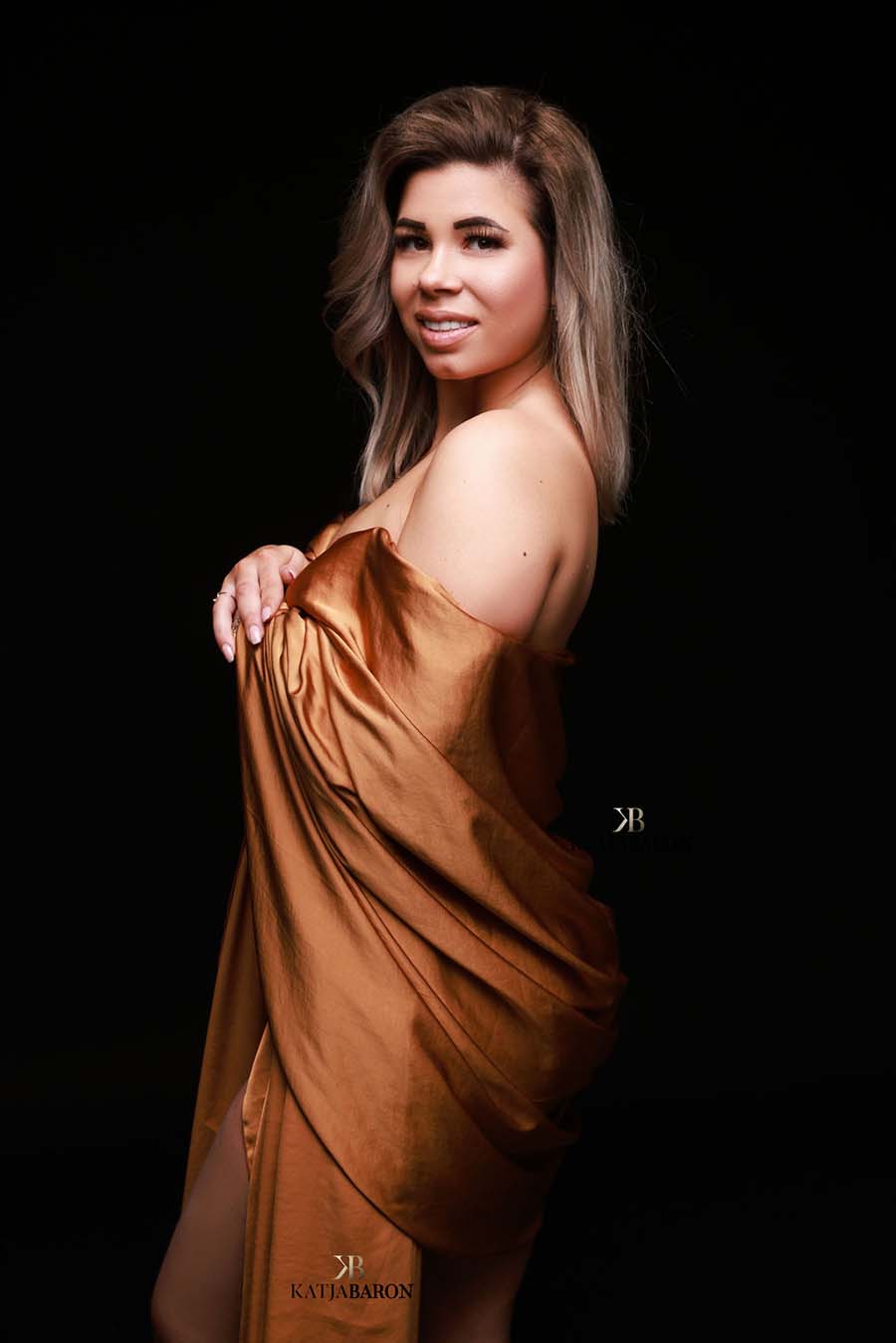 Model poses wearing a cognac camel scarf wrapped around her arms. She poses on her side, but stares at the camera.