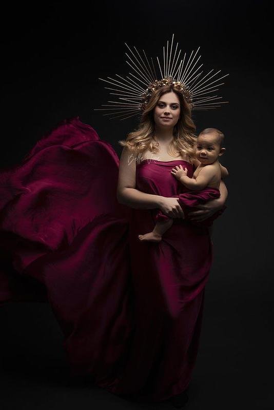 A model with blonde hair is posing with her baby boy. They are both wrapped in the same piece of fabric. The fabric is silky and in the color cherry.