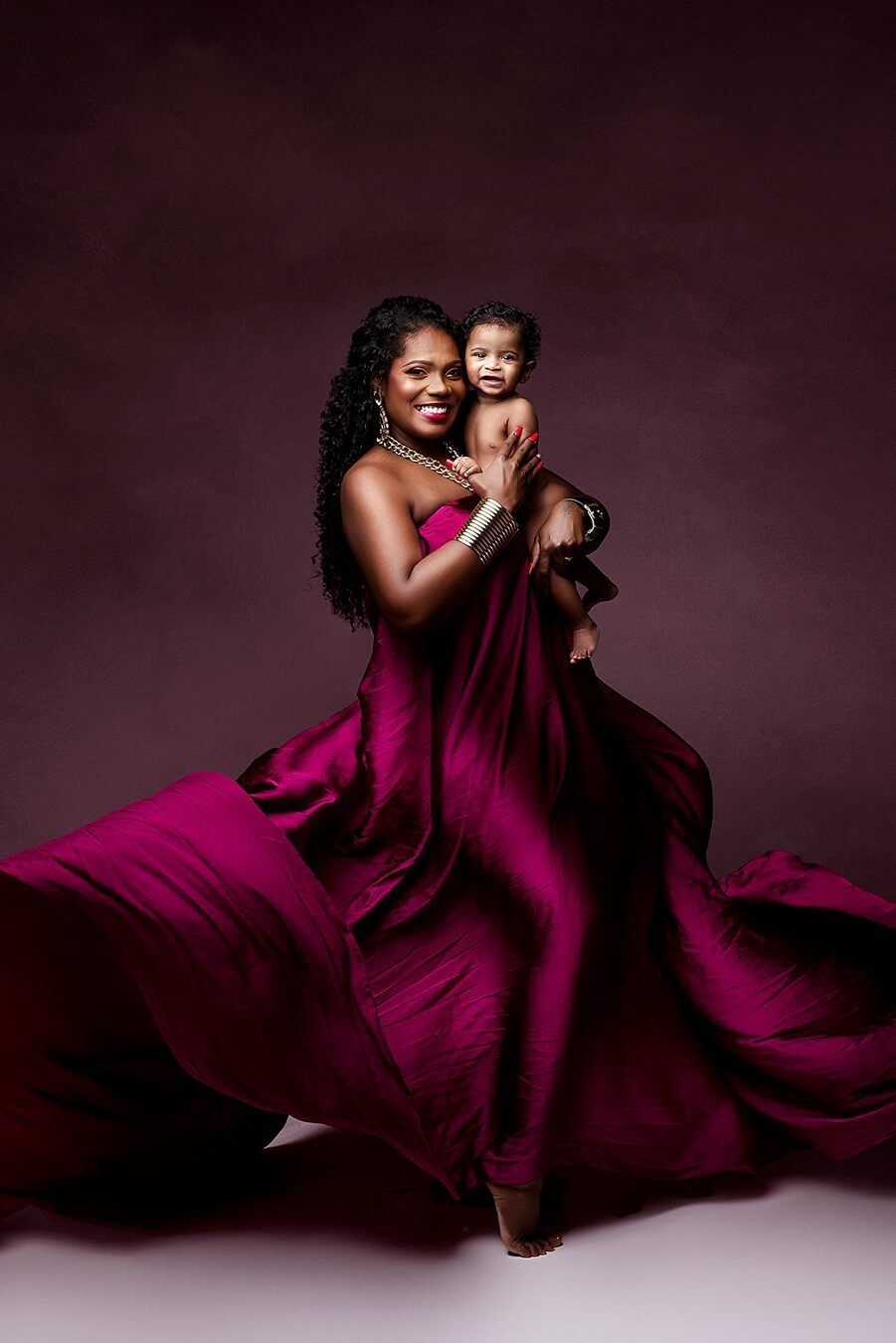 A mother is holding her baby boy. The woman is wearing a dress that is made out of scarf fabric. The color is cherry and the fabric is silky for a shimmer effect. The dress is being tossed around her for a big effect.