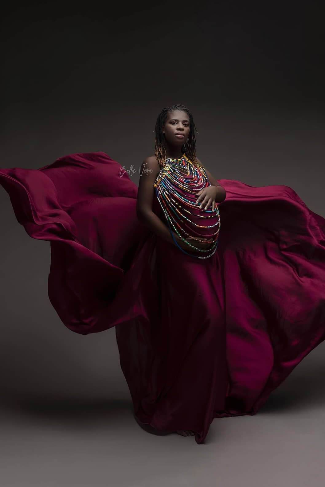 A pregnant woman is wearing a silky scarf cherry as a dress. The dress is floating around her for a big effect. She is wearing a big necklace with lots of color. The necklace has lots of layers.