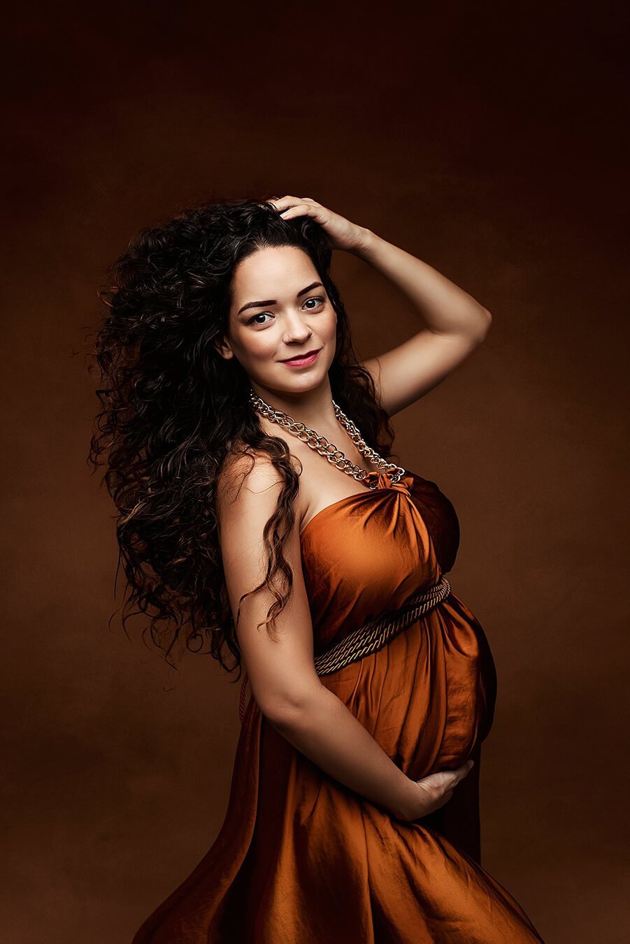 Pregnant model has curly hair and poses on her side with a scarf tied in a necklace. the whole composition looks like a dress. the scarf is made of silk and has a cognac color.