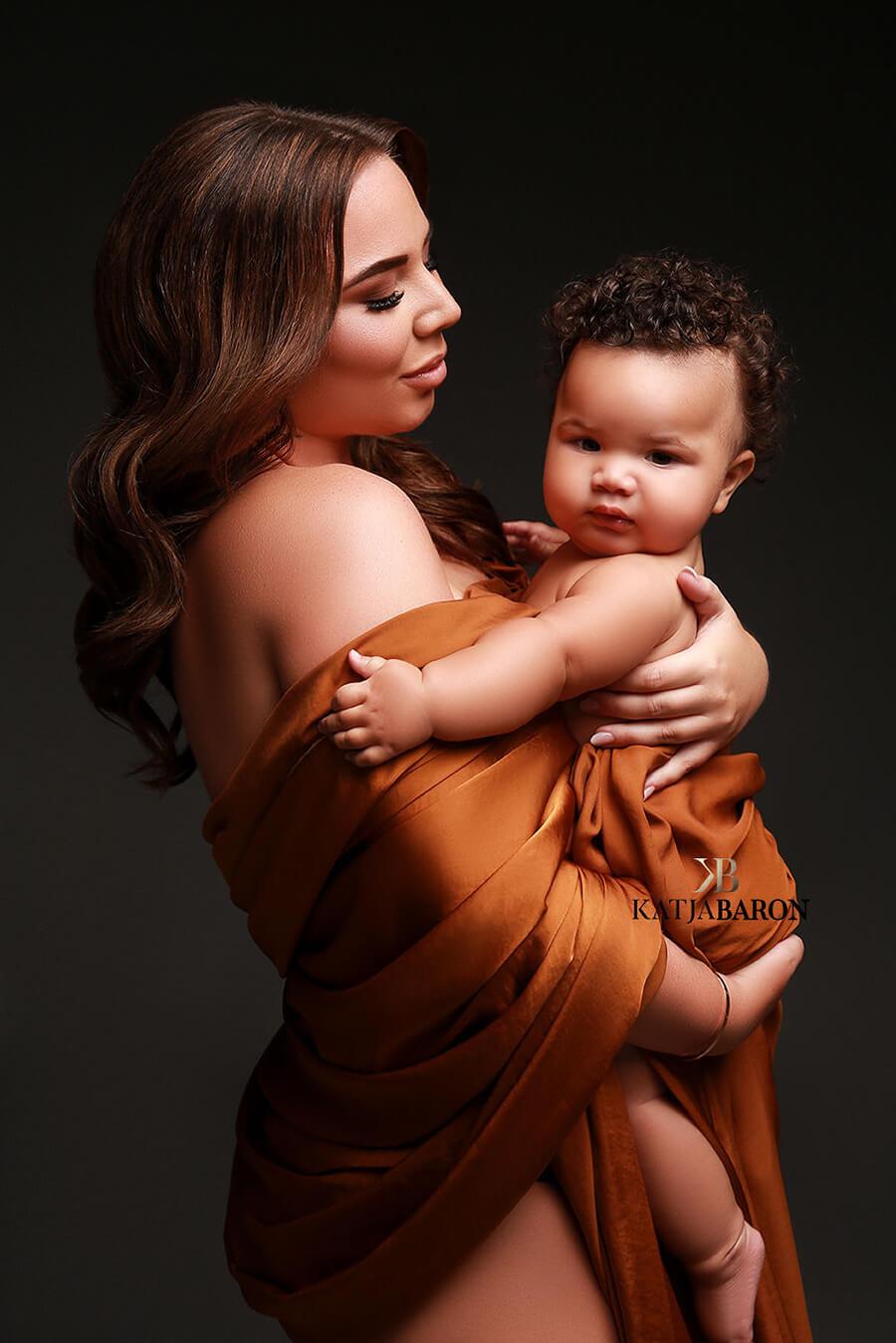 Model poses in a studio together with her baby. She covers herself and the kid with a cognac silky scarf. She is looking to her baby and the baby looking to the camera.
