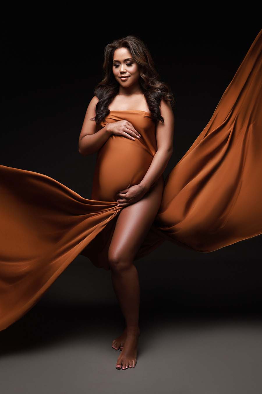 Brunette model poses wearing a cognac camel silky fabric. She looks down and holds the fabric to cover her belly and chest.