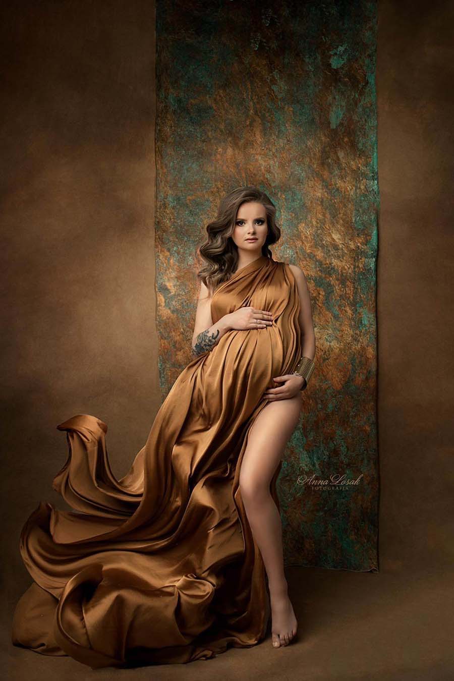 Model poses in a studio wearing a cognac camel silky scarf. Her arms and legs are uncovered and she is staring at the camera while holding her bump.