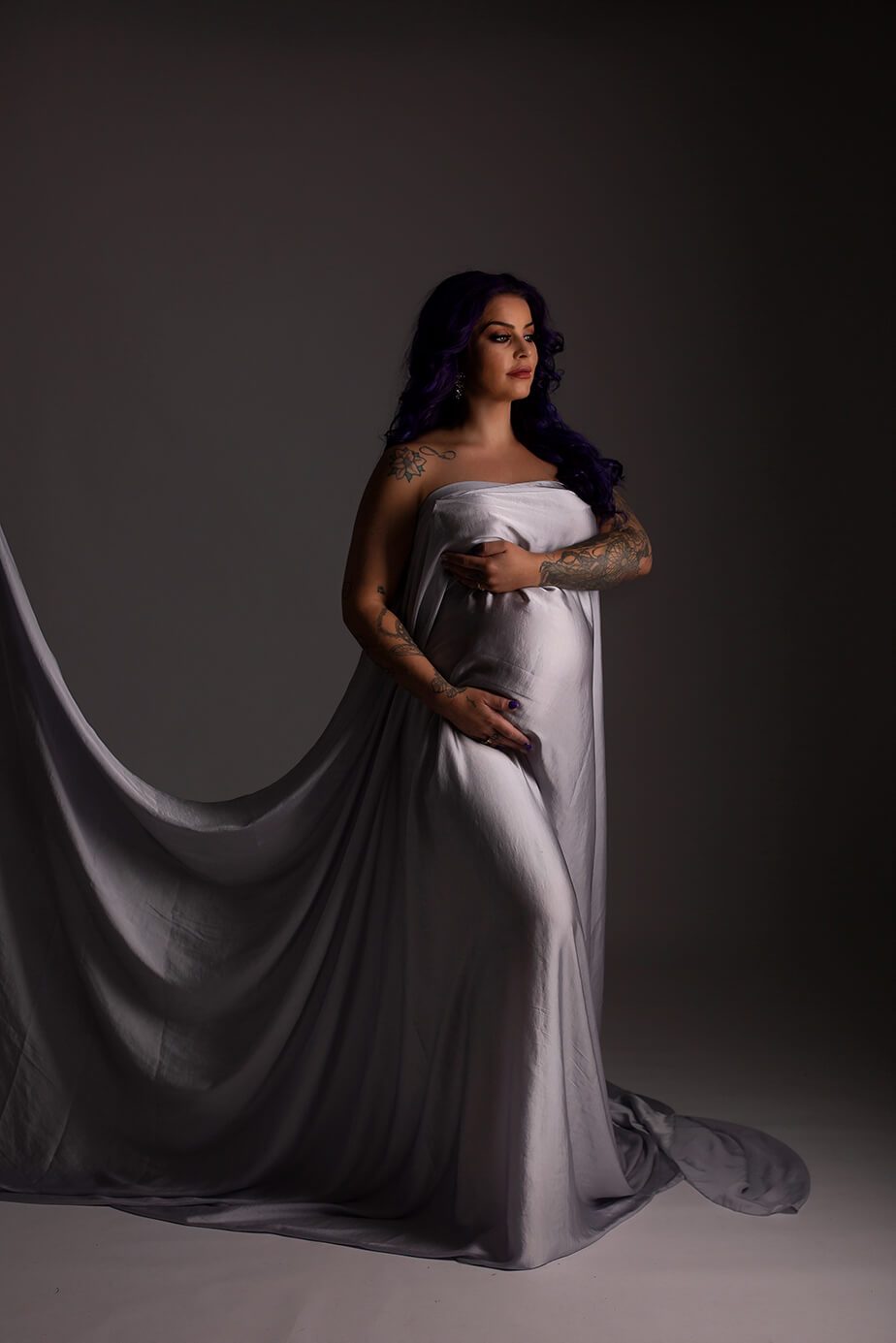 A woman is looking towards the light. She is pregnant and has purple hair. The scarf is being hold up in a curve behind her.