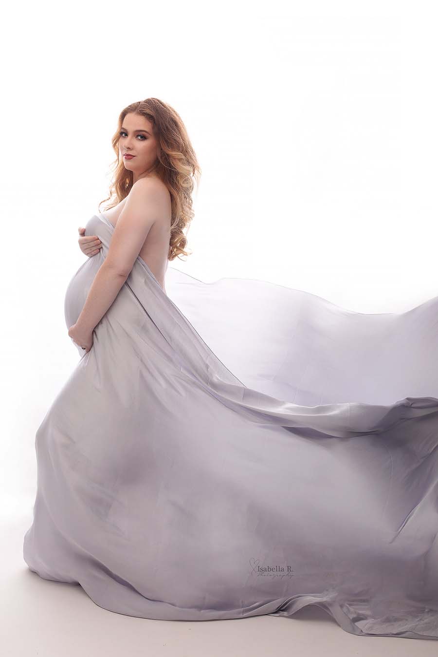 A pregnant woman is holding a scarf. the scarf is at the front of her and is going around her till the back of the photo. There is a wave in the fabric behind her.