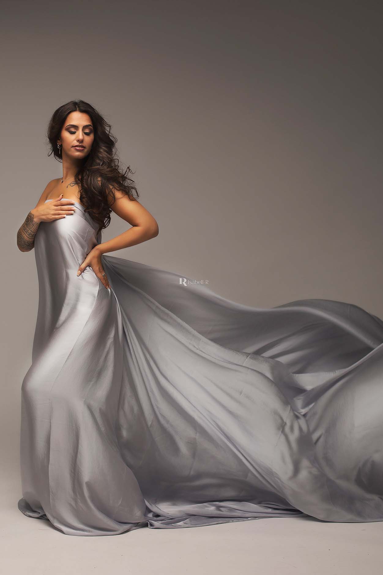 A model with long brown hair is posing in a studio. She is looking down over her shoulder. The fabric is waving behind her out of the photo.