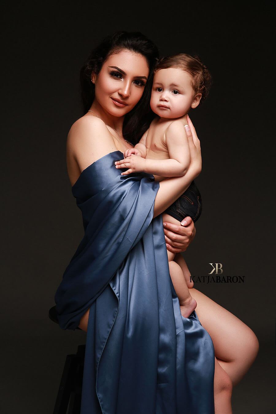 mother poses together with her baby boy in a studio. the boy has a small jeans shorts and looks to the front, while the mother has a piece of denim draping fabric around her arms. the mother is facing the camera while holding her boy.