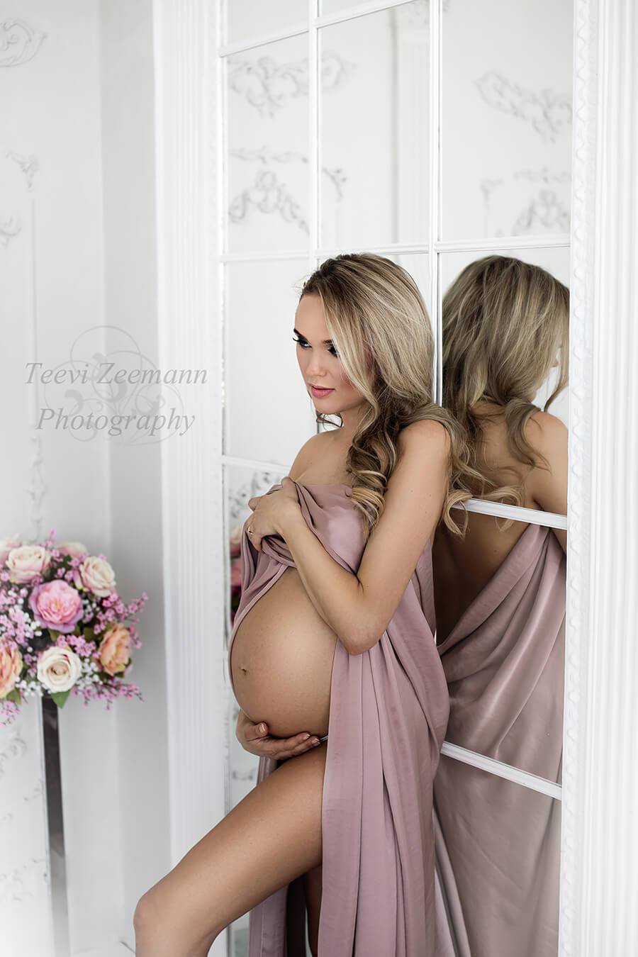 Blond pregnant woman poses against a mirror wall covering her breast with a piece of dusty pink silky fabric. She is staring to the other side and has a flower bouquet next to her.