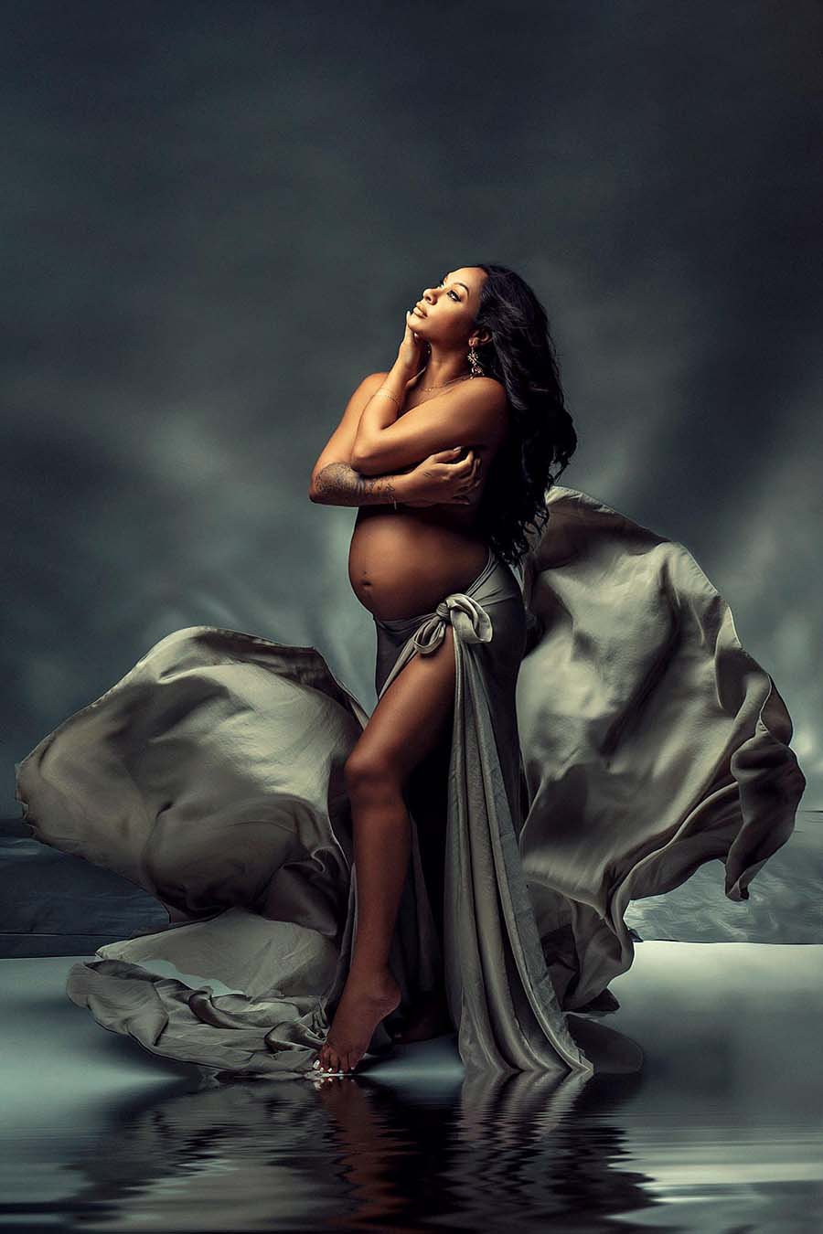 model poses by the water wearing a khaki silky scarf tied as a skirt. she is holding herself and her belly is uncovered.