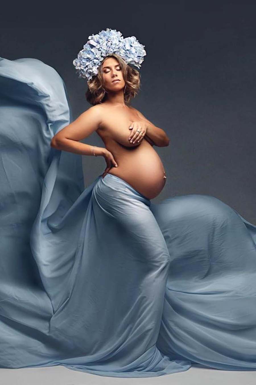 Blond short haired model poses in a studio during a maternity photoshoot. She wears only a scarf tied on her hips. The scarf is playful flying while the model has her eyes closed. She covers her breasts with one hand and holds her back with the other one. The model has a flower crown in her head.
