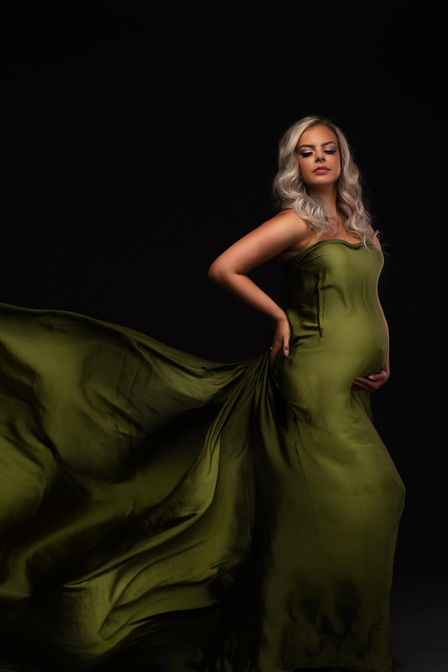 Blond Pregnant model wears a silky moss green draping fabric around her body during a maternity photoshoot. She poses on her side while holding her bump and waist. Her eyes are partially closed.