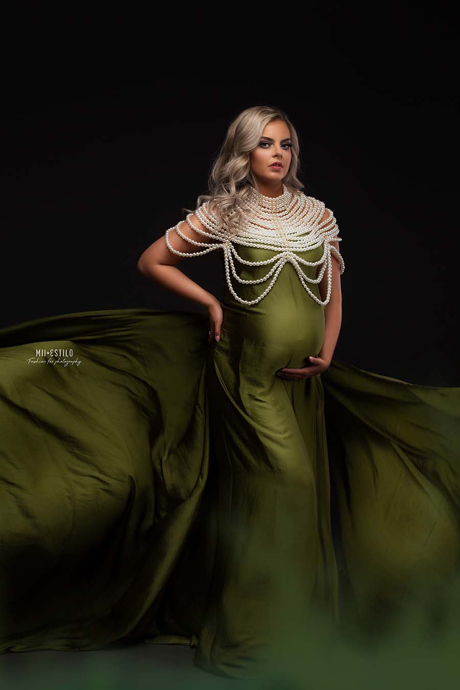 A blonde model is posing for the camera. She is wearing a silky scarf moss green as a dress. The sides are going out of the photography. She is wearing a big white pearl necklace on top of it.