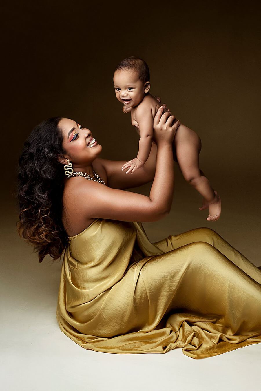A mother is holding up her baby. she is sitting on te ground and smiling towards her baby. She has the silky scarf in the color mustard around her.