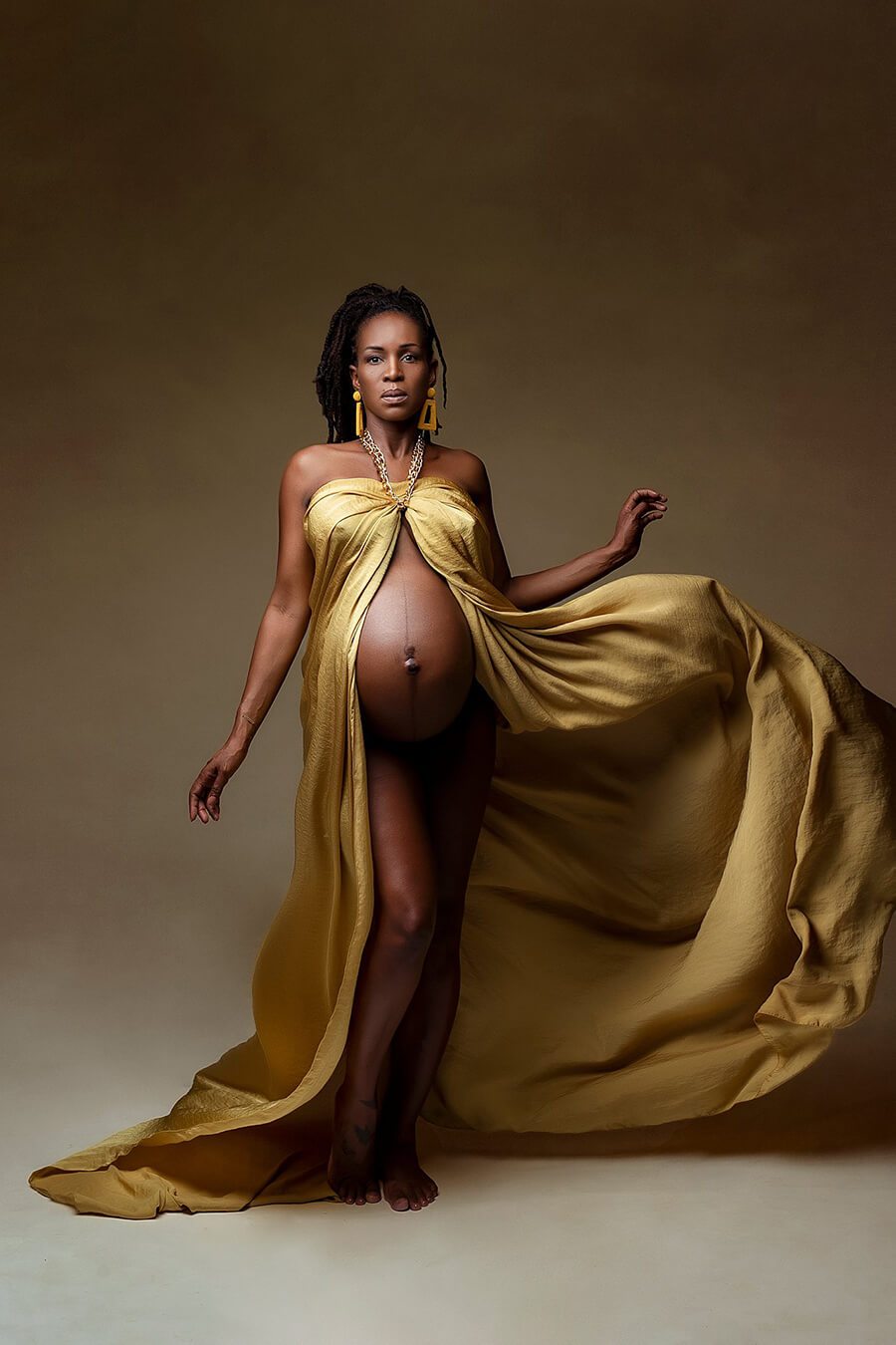 A pregnant model is wearing a silky scarf mustard. The fabric has a shimmer look. The scarf is wrapped like a dress and open at the front. You can see her babybelly. She has her hair in a ponytail. She has tossed the fabric so its stil in the air and gives the photo an extra effect.