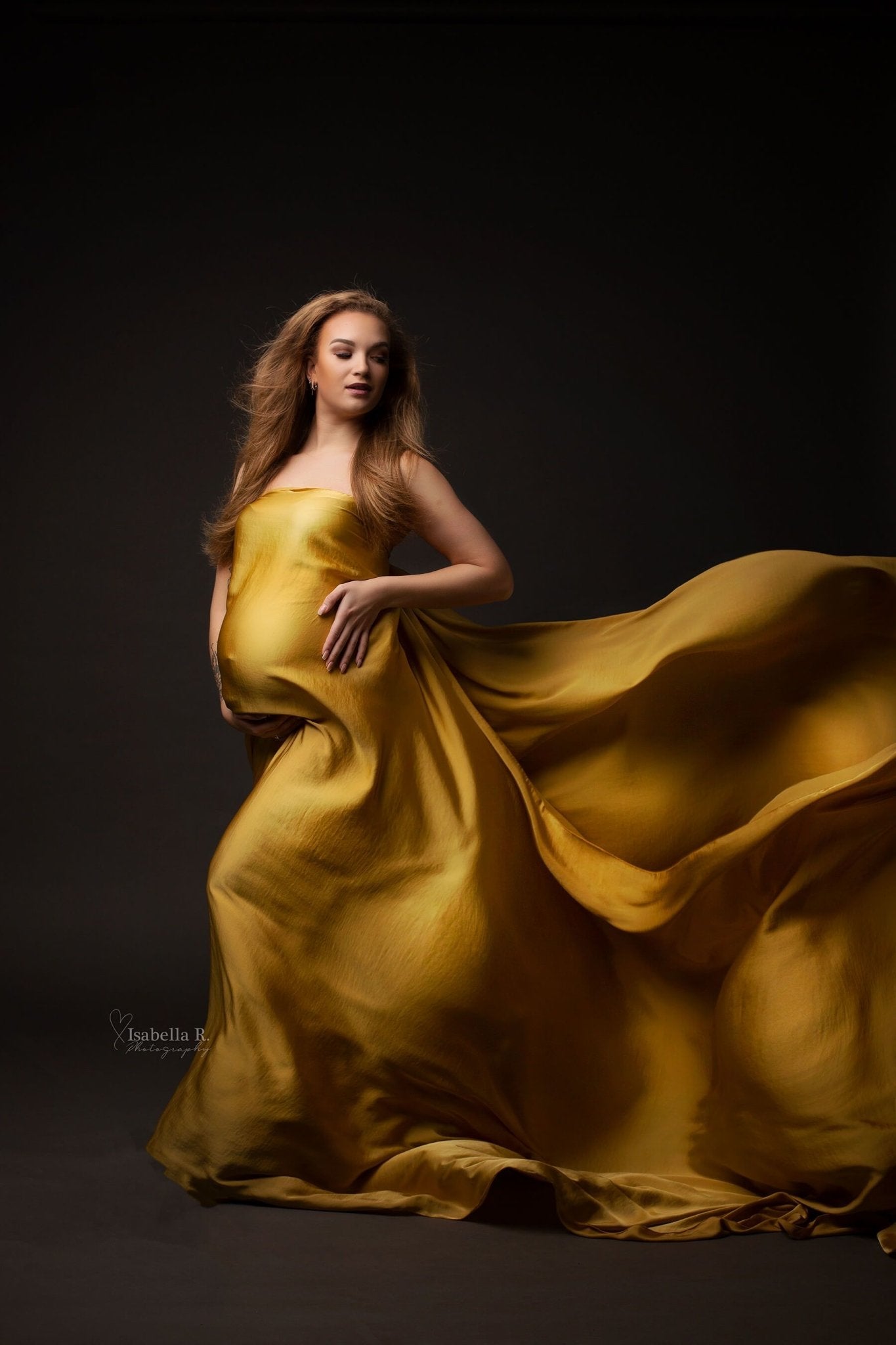 A pregnant model is standing in a studio. The fabric of the silky scarf is around her and goes to the back till the end of the photo. The fabric behind her makes waves. The model is posing with her hands underneath her belly. She is looking over her shoulder.