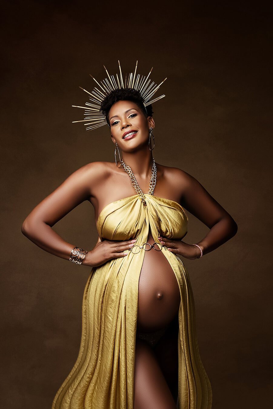 This pregnant model is smiling to the camera. she is wearing a big golden grown and other gold accessoires. The scarf is tied around the necklace and is hanging down from that point.