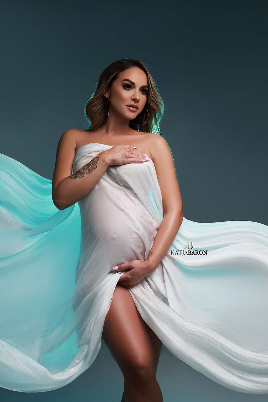 pregnant model poses with a blue background wearing a scarf in off white to cover her body.