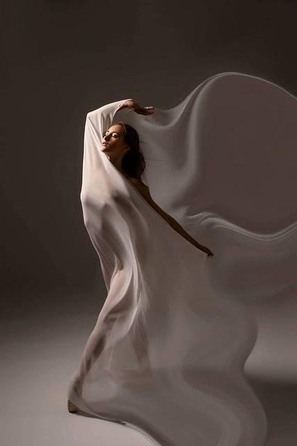 ballerina poses holding a long silky off white scarf against her body. her silhouette can can be seen through the piece.