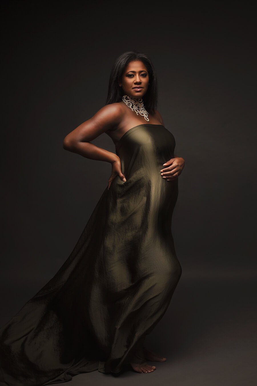 A pregnant model is looking to the camera. She has short black hair. The scarf is around her to cover her up. The scarf is in the color olive and has a silky material for a little bit of shimmer. The woman is wearing a big white necklace.