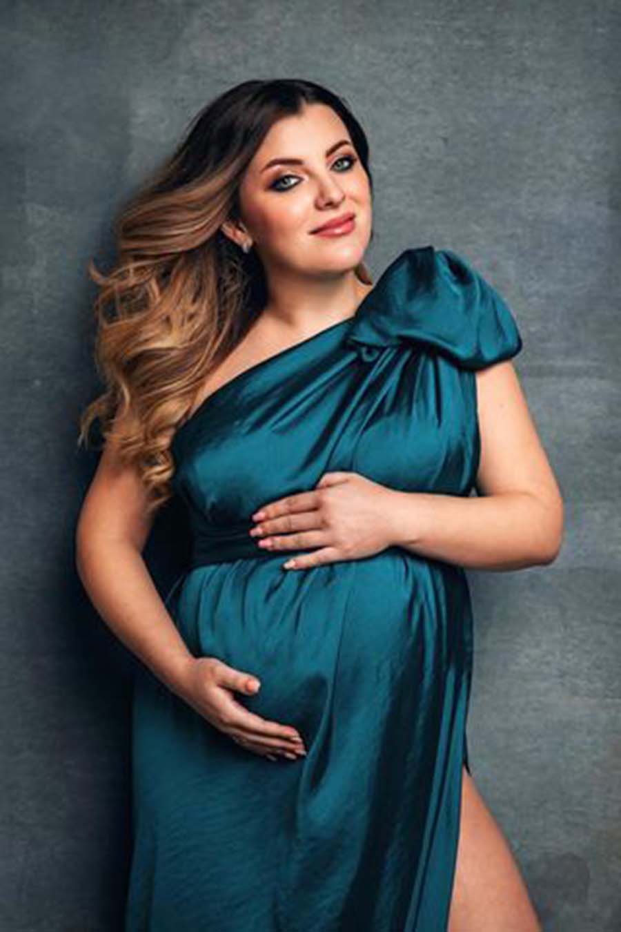 A pregnant woman has a silky scarf petrol around her. The scarf is used to create a dress. They tied a little bow on one shoulder. she is posing with her hands around her belly and smiling to the camera.