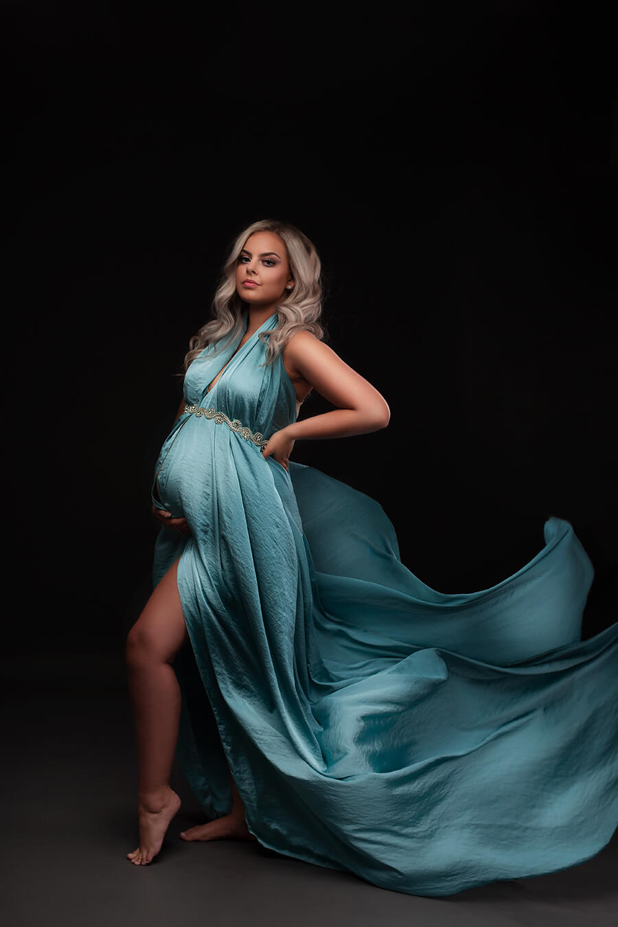Blond pregnant model wears the mii-estilo 4 meter long silky scarf in powder blue color. The scarf is dressed on the model and adorned with a sash as accessory. The model faces the camera with a side look. One hand holds her bump and the other holds her back.