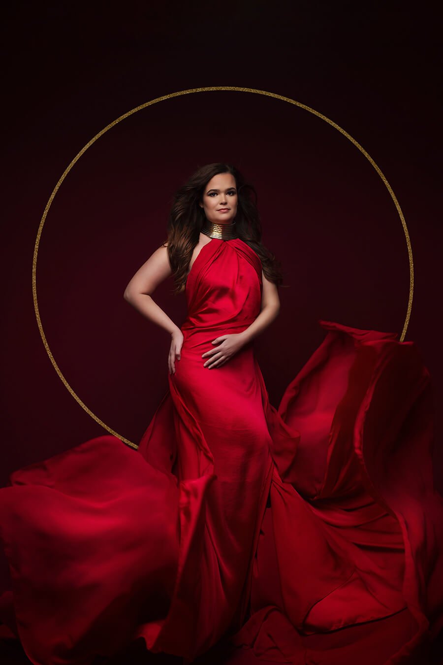 model posed wearing a long red silky scarf and a golden necklace. The scarf is attached to the body like a dress. Model stares to the camera.