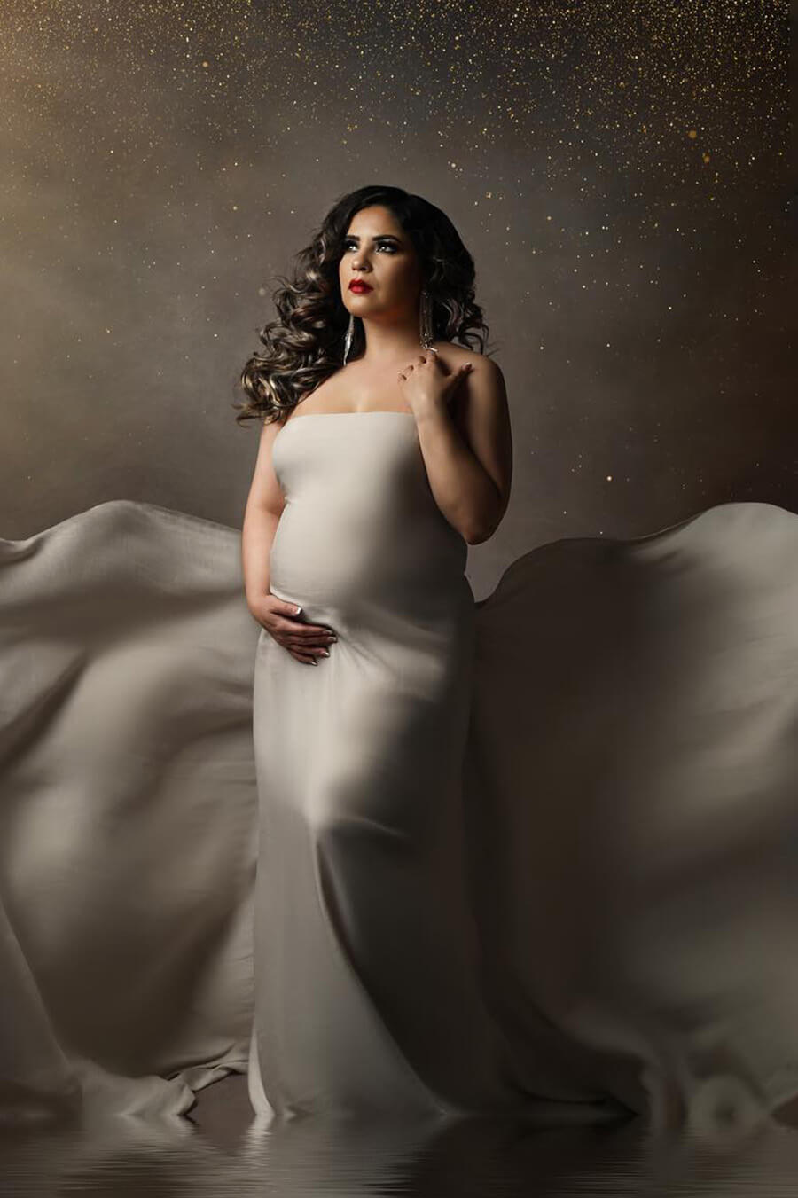 pregnant model with dark blond hair poses in a studio wearing a silky scarf in sand color wrapped around her body and with enough fabric on the back to create flying movements.
