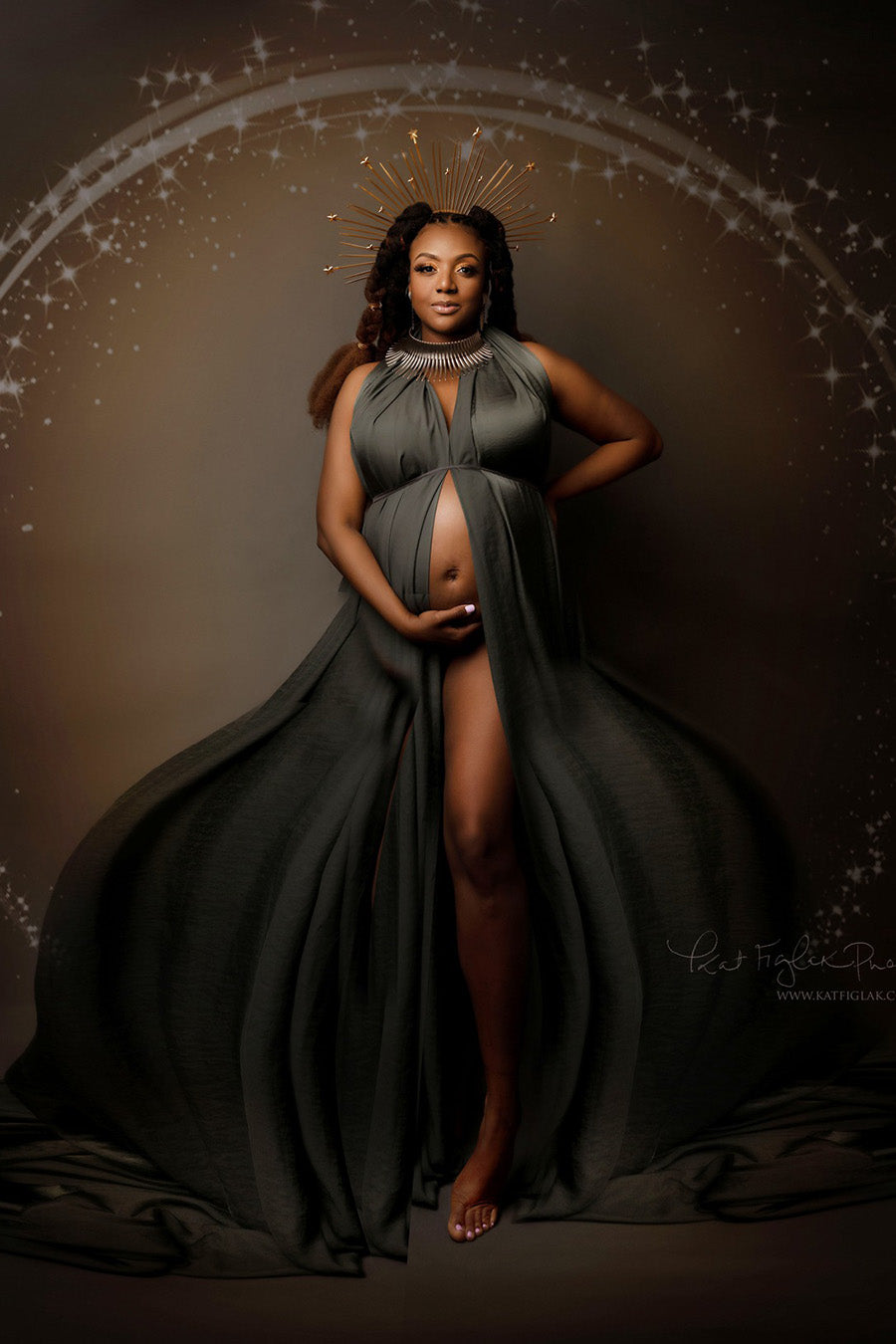 pregnant model poses against a brown background with stars. she is wearing a slategrey silky scarf wrapped around her body as a dress. she has a crown and a big necklace to match the style.