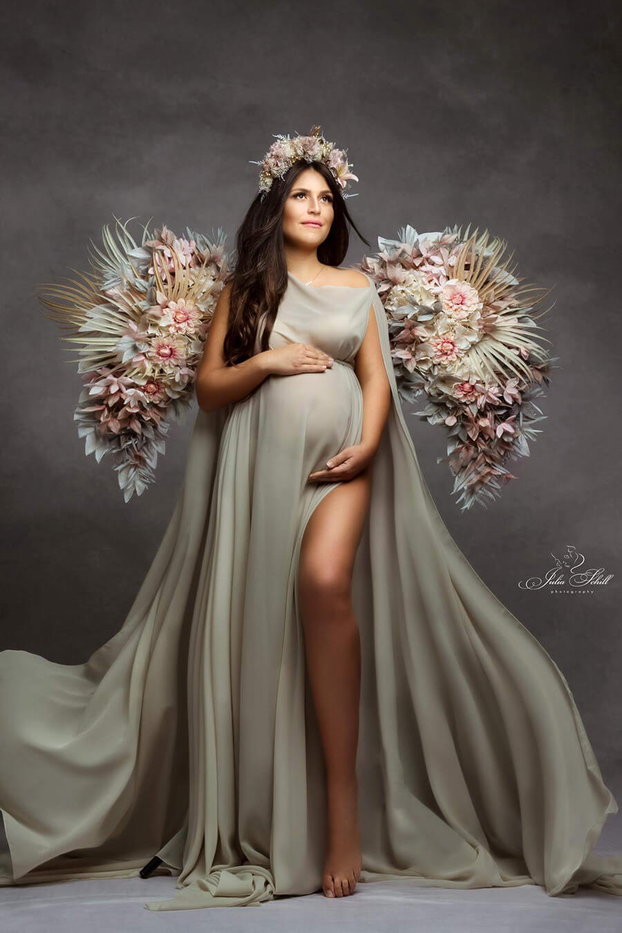 A model is posing in a studio in front of a grey background. The model has long dark brown hair. She is wearing flower wings and a matching flower headband. She is also wearing a simple but classy chiffon cape in the color sand. The cape is long and kind of looks like a loose fitted dress. The photograph is just magical.