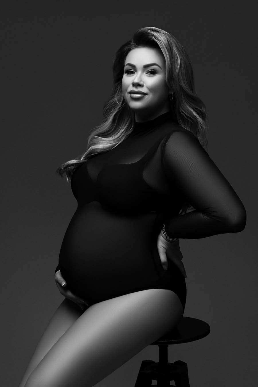 pregnant model is sitting in a stool while posing for a maternity photoshoot. she is wearing a transparent bodysuit made of mesh with a bra underneath. she is facing the camera and holding her back and belly with her hands. her hair is long and blond and the photo is black and white.