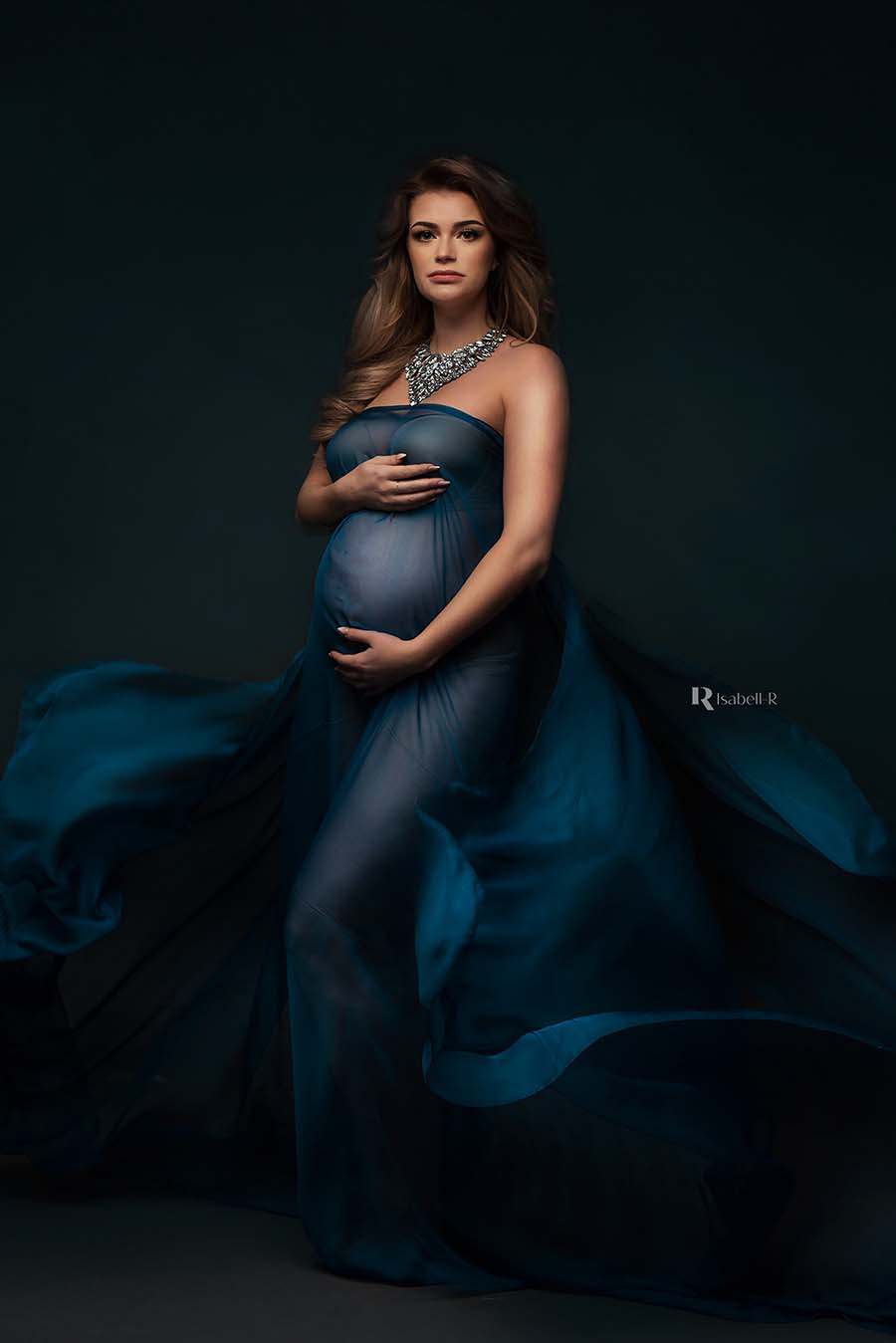 A pregnant model is wearing a voile scarf as a dress. The color of the scarf is petrol and the fabric is a little bit sheer. She is wearing a big statement necklace to finish the look.