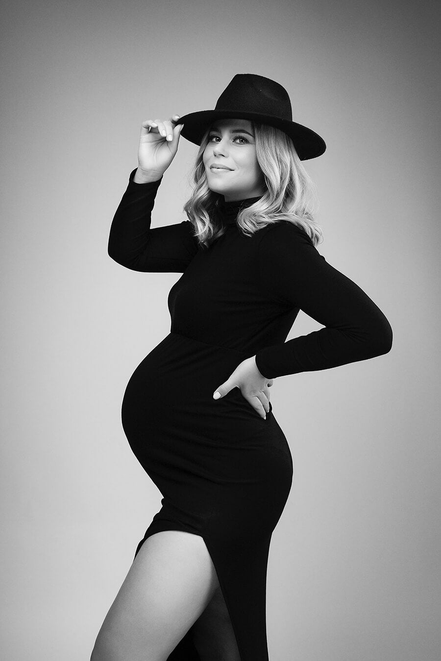 black and white photo of a pregnant model wearing a tight long black dress with a split on the skirt. the dress features a turtleneck neckline and long sleeves. the model wears a hat to match the look. she is posing on her side and has one of her hands holding the hat and the other resting on her side.