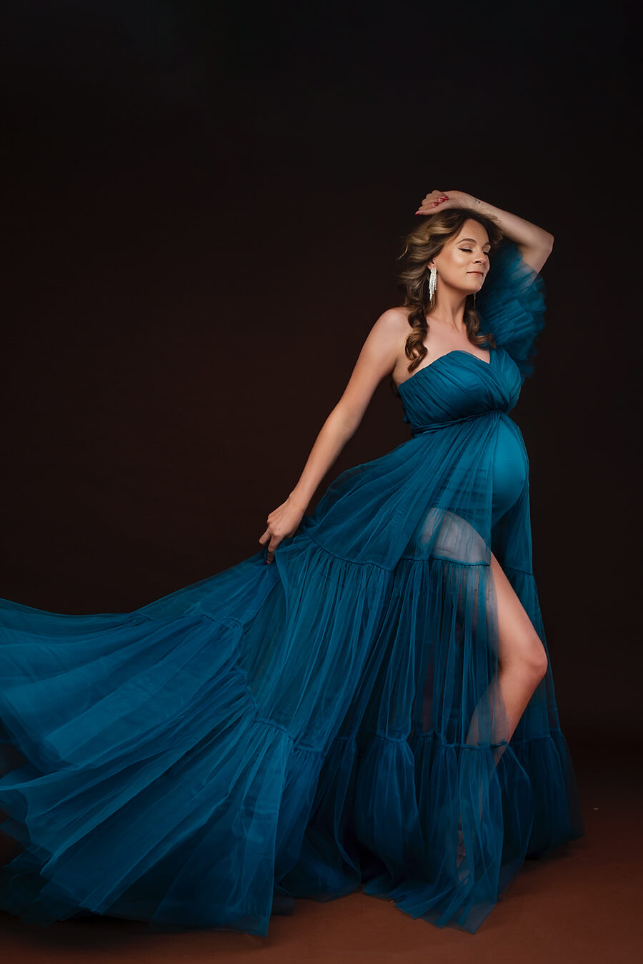 pregnant brunette model poses in a studio wearing a long tulle dress in petrol color. the model has her eyes closed and has one of her hands above her head and the other holds the skirt from the dress. the tulle dress has a bodysuit under and a middle split on the skirt. This one shoulder dress has lovely tulle details on one of the sleeves and a sweetheart neckline to finish it up.