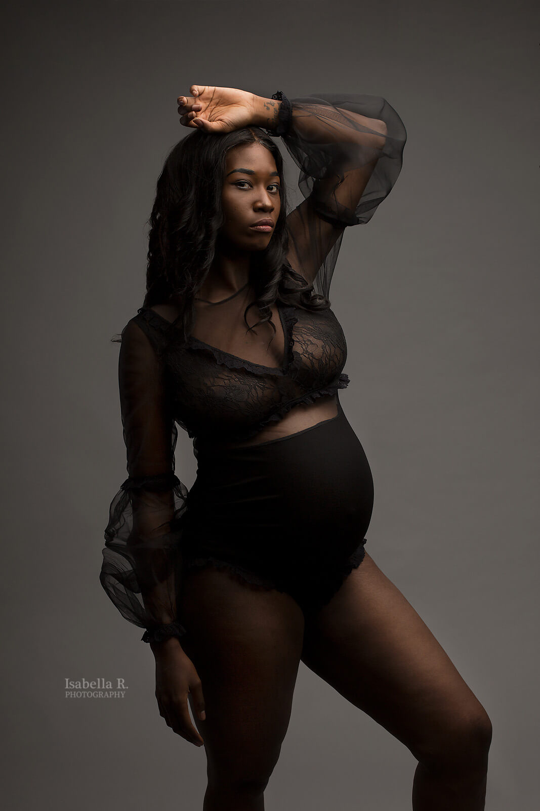 The woman in this picture has black short hair and is pregnant. She is posing with one hand above her head and the other hand is hanging next to her. The bodysuit is sheer and black. The bottom half is made from jersey and isn&