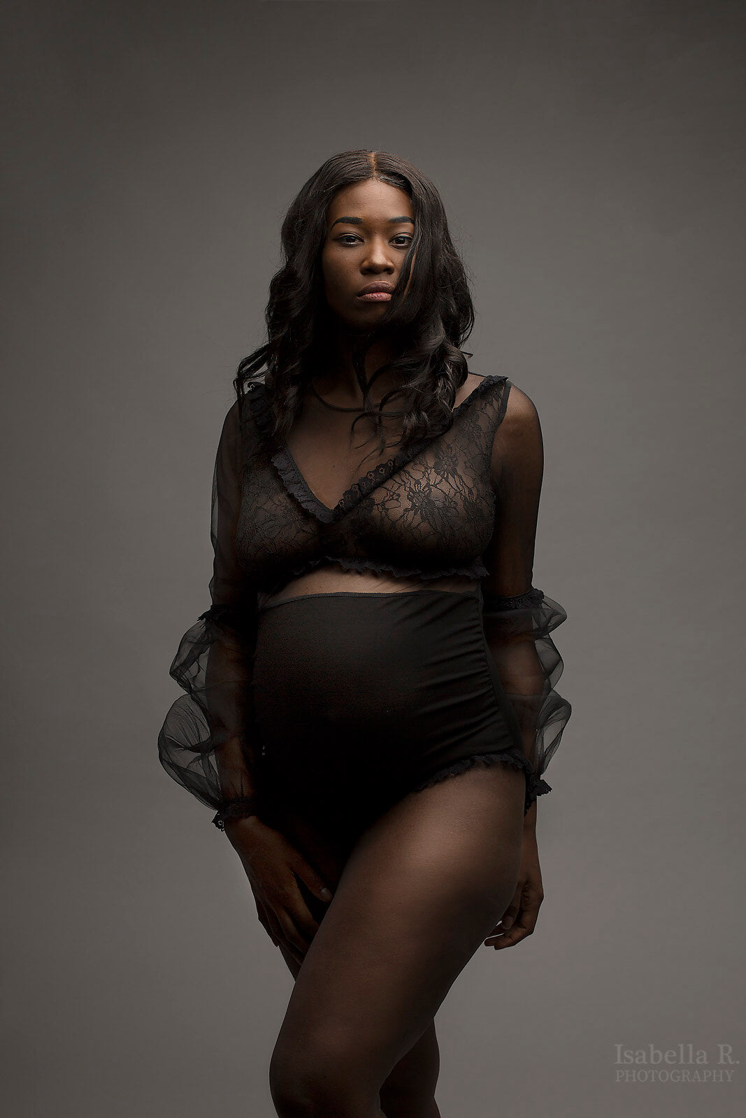 The model has black curly hair. The model is wearing a black sheer bodysuit. The top is made from lace and the bottom is a black jersey material. The model is posing with one leg in front of the other leg and looking into the camera. 