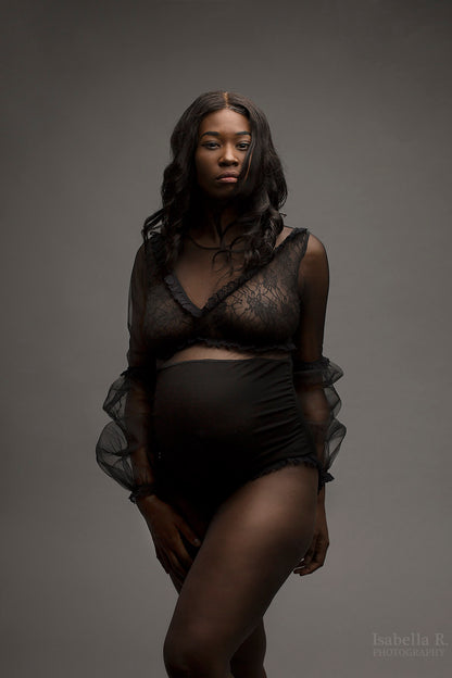The model has black curly hair. The model is wearing a black sheer bodysuit. The top is made from lace and the bottom is a black jersey material. The model is posing with one leg in front of the other leg and looking into the camera. 