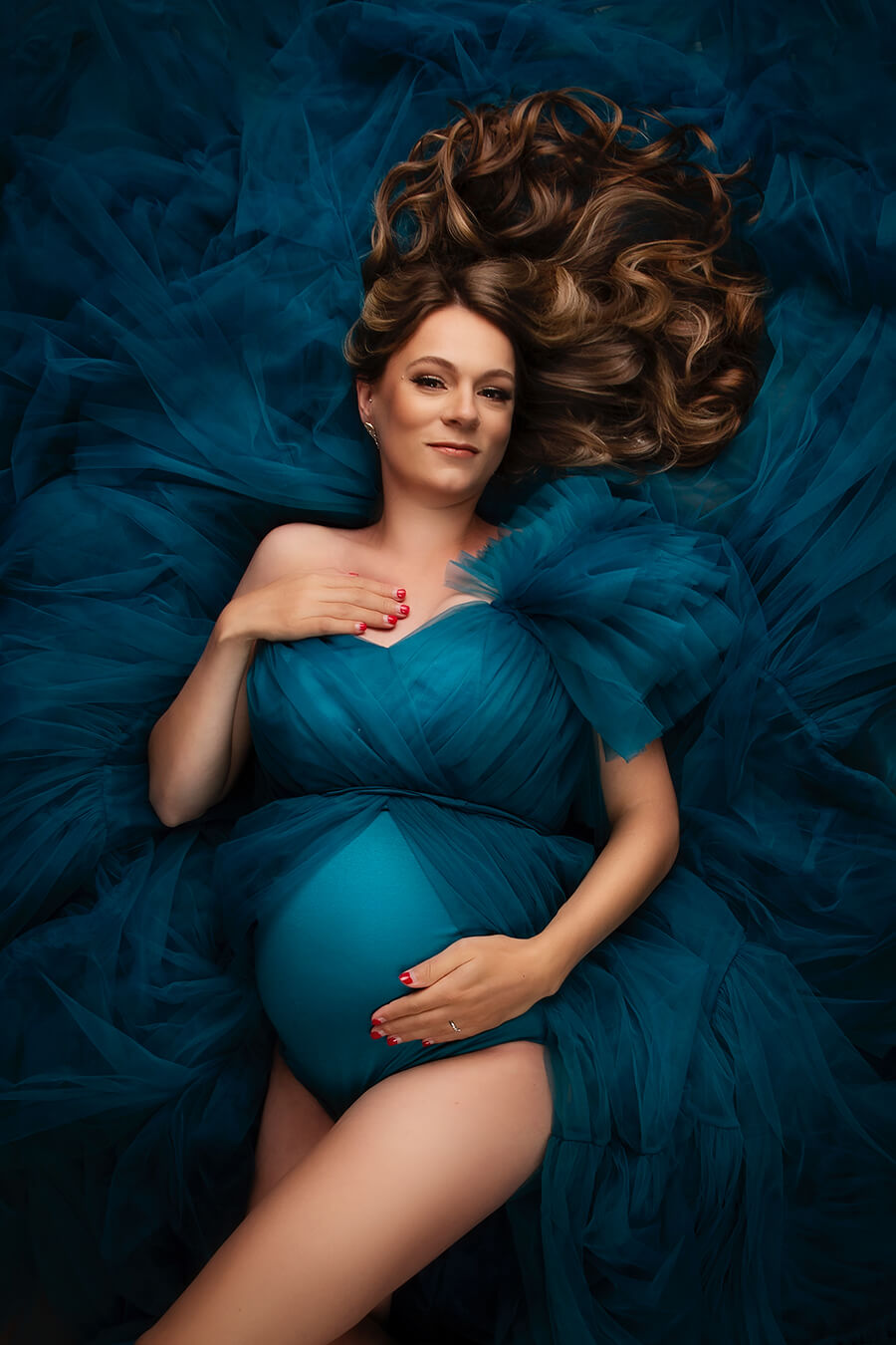 pregnant brunette model lays on the studio floor on the excess of tulle fabric. she is wearing a tulle dress in petrol color with a bodysuit under.