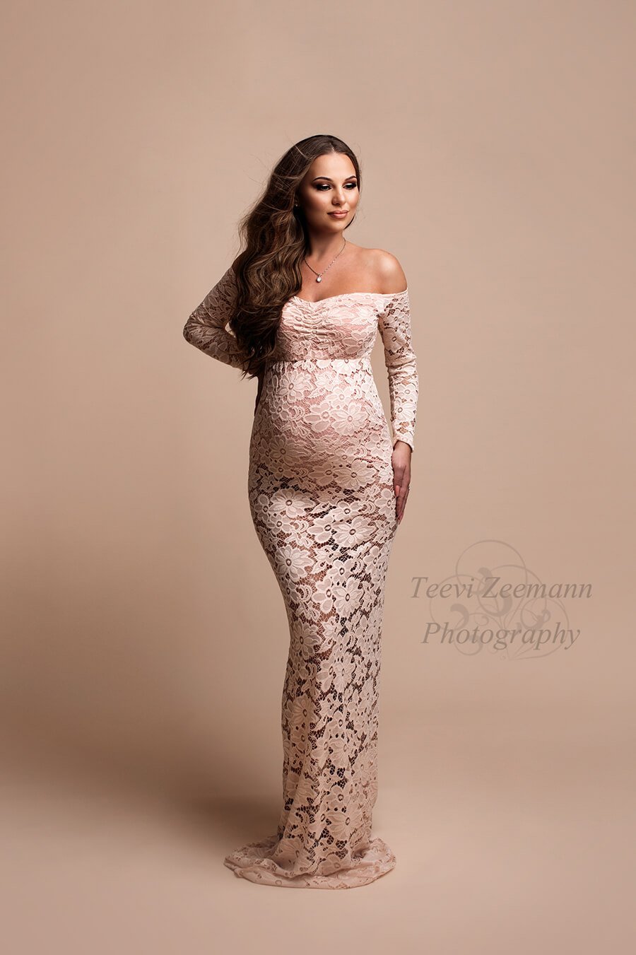 In the studio is the model wearing a long dress made out of lace. The dress is see trough at the skirt. She is pregnant