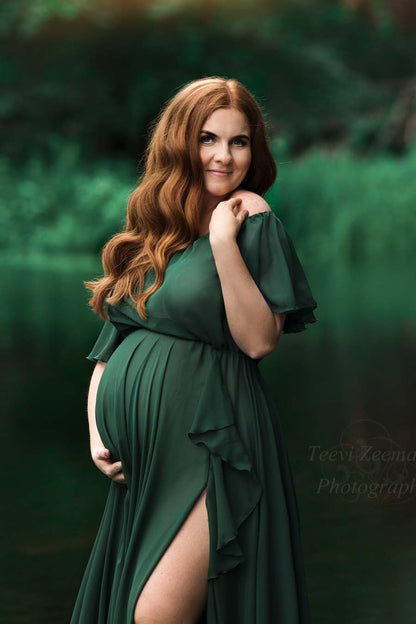 red haired pregnant model poses wearing a long dark green dress made of chiffon. the dress features an off the shoulder top with ruffle short sleeves. the skirt has a split on the side with ruffle details at the endings. 