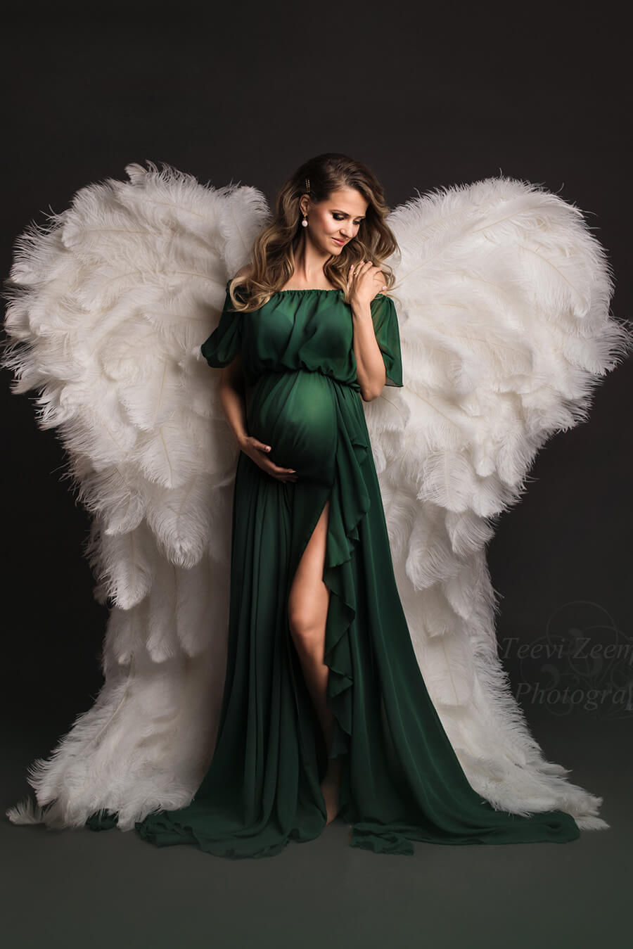 pregnant model poses in a studio wearing a dark green chiffon dress with ruffle details and a side split. she is wearing a fake big wing on her back in white color. 